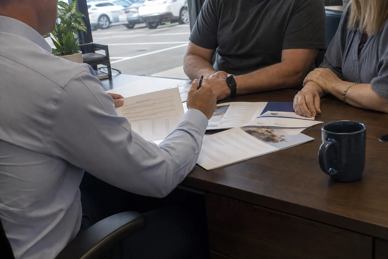 Agent in a light blue button-down reviewing printed papers with a couple at a wooden desk by a window, a navy coffee mug on the desk and parked cars outside