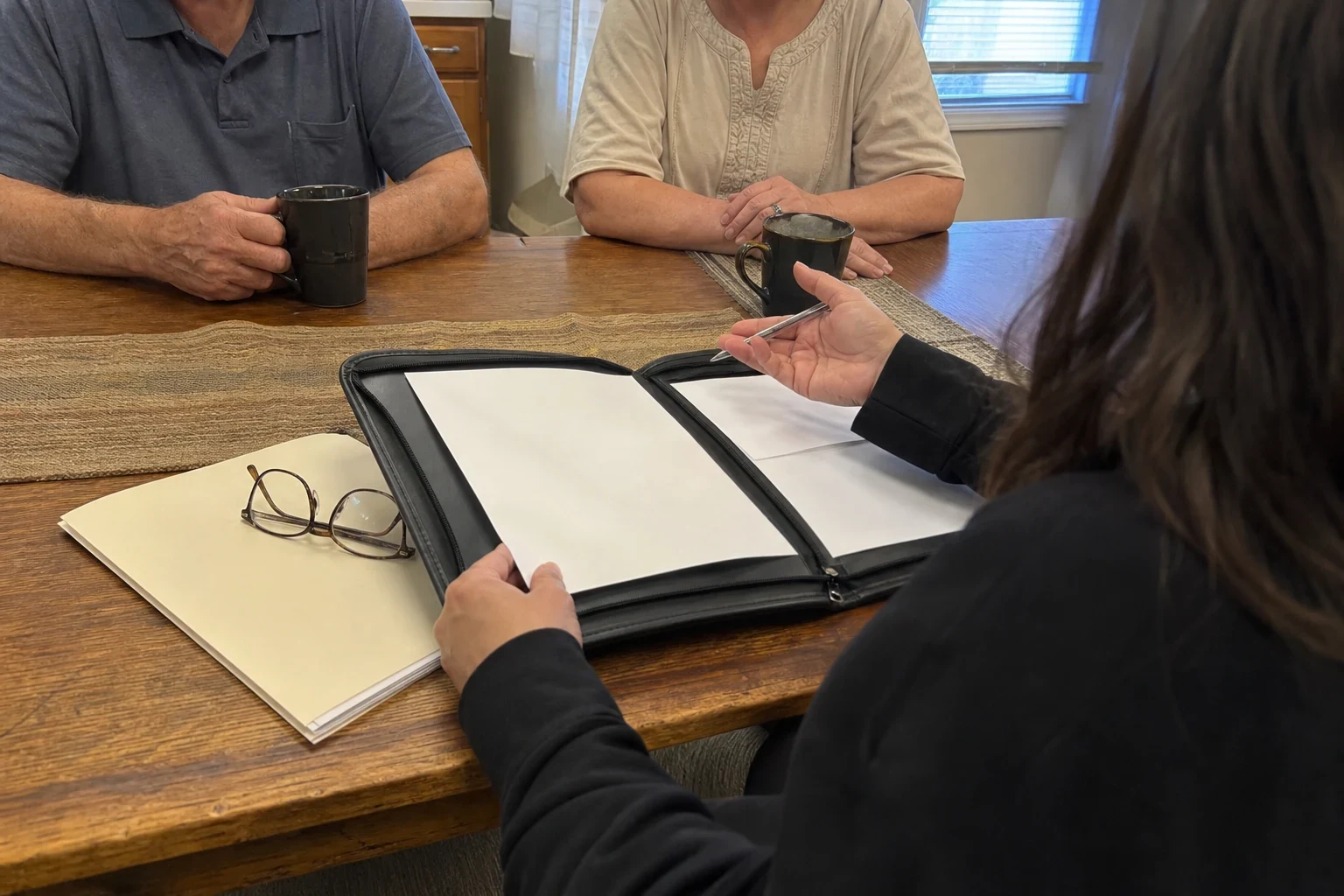 Agent with an open black portfolio and pen sits across a wooden kitchen table from an older couple holding coffee mugs, with a folder and reading glasses nearby