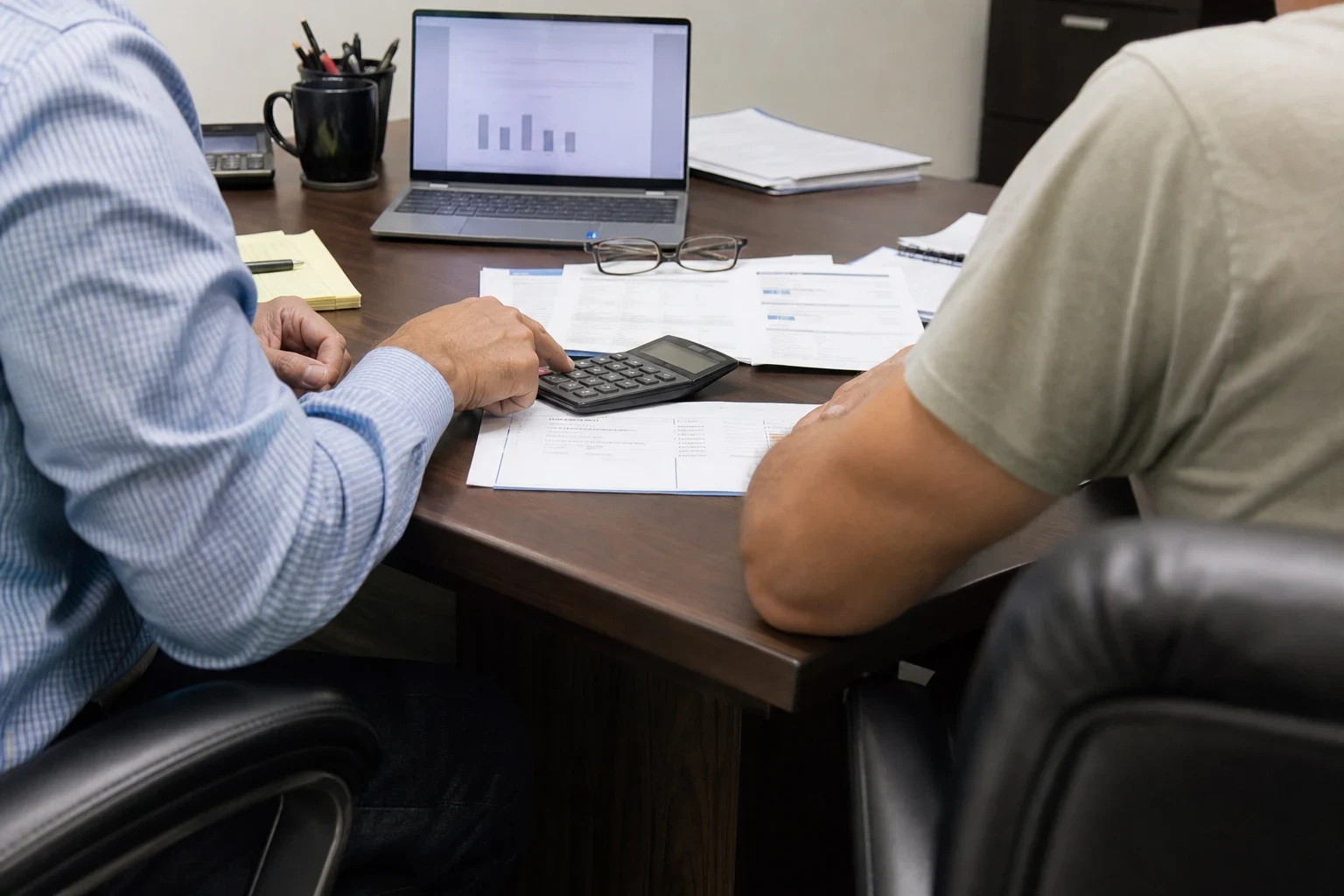 Two men seated at a dark wood office desk, one tapping a calculator on printed statements, with a laptop, notepad, glasses and coffee mug nearby