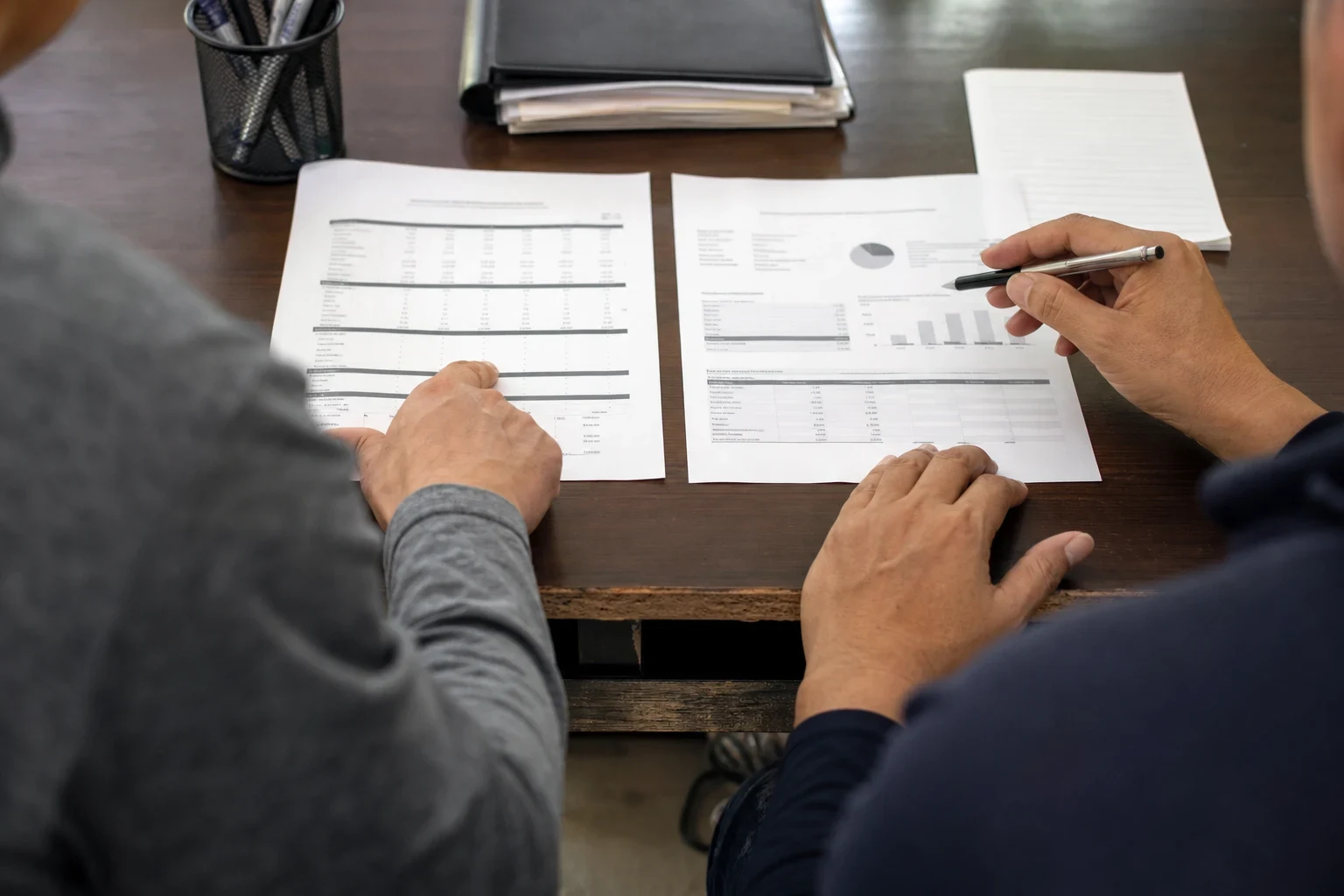 Over-the-shoulder view of two people at a wooden desk reviewing two printed documents side by side, one holding a pen, with a pen cup and folders nearby