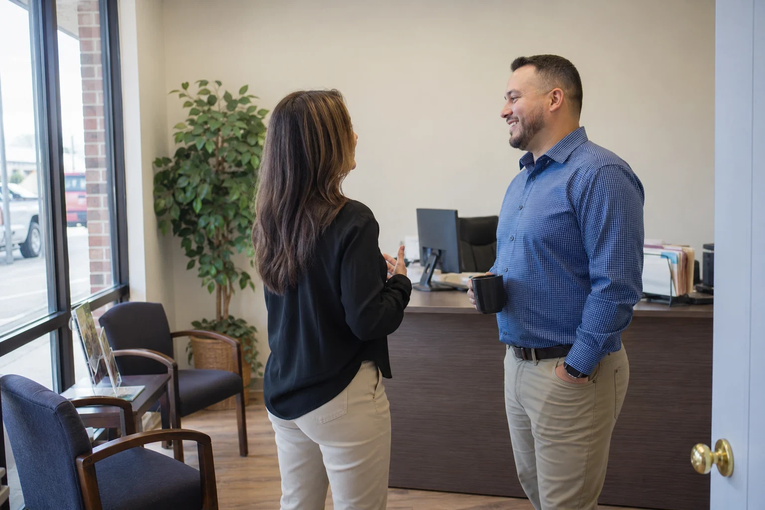Two agents chat beside a reception desk in a small office with a potted plant, waiting chairs, a brochure stand and a large street-facing window.